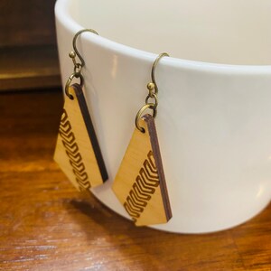 May include: A pair of wooden earrings with a geometric design. The earrings are a light brown colour and have a brass hook.