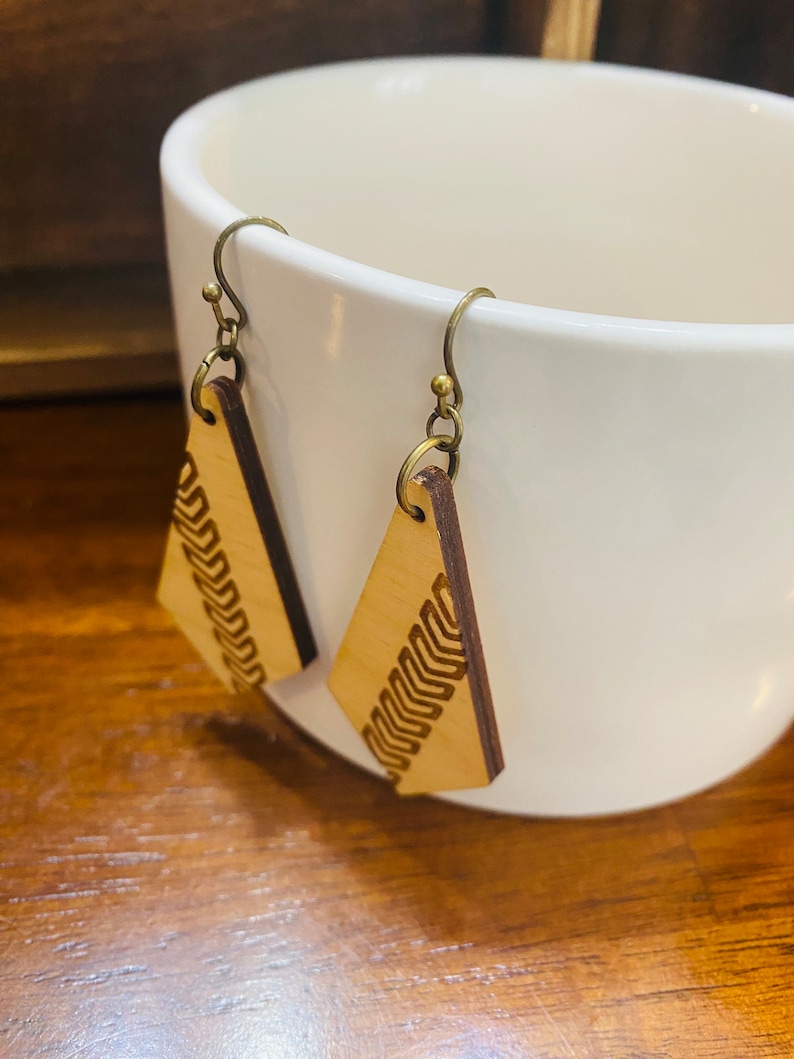 May include: A pair of wooden earrings with a geometric design. The earrings are a light brown colour and have a brass hook.