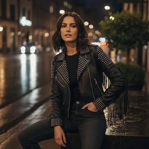 Puede incluir: Chaqueta de cuero negra con tachuelas plateadas en los hombros y el cuello. La chaqueta tiene flecos en las mangas y se combina con vaqueros negros. La imagen está ambientada en una calle de la ciudad con luces borrosas.