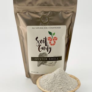 May include: A gold-colored bag of "Soil Candy" oyster shell soil conditioner. The label reads "All Natural Soil Conditioner" and "Oyster Shell." A small wooden bowl contains a pile of the gray oyster shell powder. Net weight is 2 lbs.