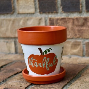 May include: A terracotta flower pot with a white base painted with a pumpkin and the word "thankful" in green lettering. The pot has a matching terracotta saucer.