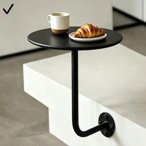 Puede incluir: Una mesa auxiliar redonda negra con una pata curva. Una taza blanca con base marrón y un plato con un cruasán están sobre la mesa. La mesa está fijada a una superficie blanca con una placa de montaje circular negra.