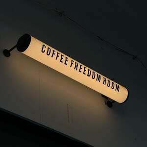 May include: A rectangular, illuminated sign with the words "COFFEE FREEDOM ROOM" in black lettering. The sign is mounted on a dark wall and has black end caps. The light is a warm, inviting color.