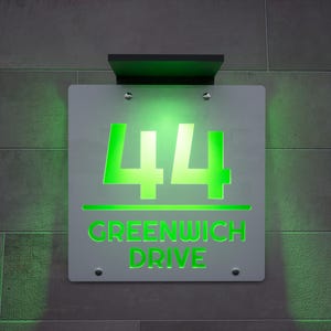 以下が含まれることがあります： 明るい緑色で「44」の数字と「GREENWICH DRIVE」の文字が表示された、照明付きの住所看板。 看板は灰色のタイルの壁に取り付けられており、黒い上部が付いています。