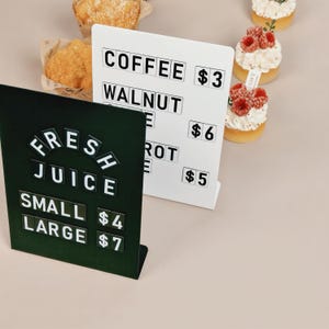 May include: A dark green metal sign displaying the text FRESH JUICE with SMALL $4 and LARGE $7, positioned in front of a white metal sign displaying COFFEE $3, WALNUT $6, and CARROT $5, accompanied by several small pastries with raspberries.
