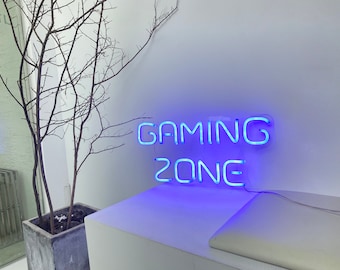 Puede incluir: Un letrero de neón azul que dice "GAMING ZONE" montado en una pared blanca. El letrero está iluminado y proyecta un brillo azul.