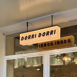 Op de afbeelding: Een rechthoekig verlicht bord met de tekst "DORRI DORRI" in vette zwarte letters. Het bord hangt aan het plafond boven een glazen winkelpui. Een tweede bord met dezelfde tekst wordt weerspiegeld in het glas.