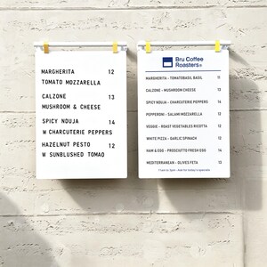 May include: Two white rectangular menu boards with black text hanging from white bars with yellow clips. The left board lists pizza options like Margherita and Calzone. The right board features the Bru Coffee Roasters logo and a list of pizzas.