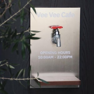 Puede incluir: Un letrero de acero inoxidable para "Vee Vee Cafe" con un grifo y horario de apertura. El grifo tiene un mango rojo. El letrero está montado en una pared de madera oscura, con hojas verdes en primer plano.
