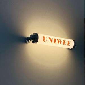 以下が含まれることがあります： オレンジ色の文字で「UNIWEE」と書かれた円筒形の照明器具。器具は壁に取り付けられており、端と取り付け金具にはクロム仕上げが施されています。ライトが点灯し、周囲の壁を照らしています。
