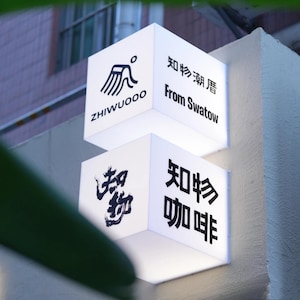 May include: Two illuminated white cube signs with black text and graphics. The top sign reads "ZHIWUOOO" and "From Swatow". The bottom sign reads "咖啡" and "知物". The signs are mounted on a light-colored wall.