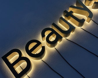 May include: A gold-colored sign with the word "Beauty" in black letters. The letters are raised and illuminated from behind, creating a glowing effect.