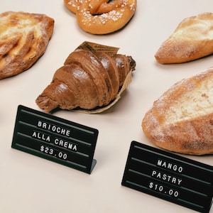 May include: A collection of assorted baked goods including a golden-brown croissant in a paper sleeve, a pretzel, and two loaves of bread. Two black rectangular signs with white lettering are present, reading BRIOCHE ALLA CREMA $23.00 and MANGO PASTRY $10.00.