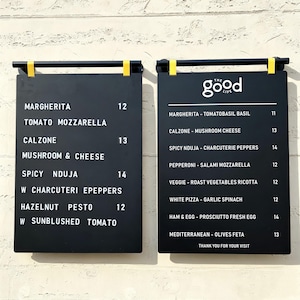 May include: Two rectangular black menu boards with white text mounted on a wall. The left board lists pizza options like Margherita, Calzone, and Spicy Nduja. The right board features the logo The Good Life and lists various pizza selections.