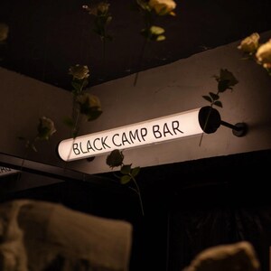 May include: A glowing, rectangular sign that reads "BLACK CAMP BAR" in white lettering. The sign is mounted on a wall and surrounded by hanging flowers. The overall aesthetic is dark and moody.