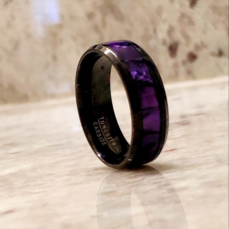 Pode incluir: Um anel de carboneto de tungst&ecirc;nio preto com uma incrusta&ccedil;&atilde;o roxa. O anel tem as palavras "TUNGSTEN CARBIDE" gravadas no interior.
