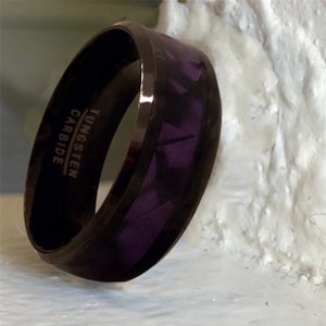 Pode incluir: Um anel de carboneto de tungst&ecirc;nio preto com uma incrusta&ccedil;&atilde;o roxa. O anel tem as palavras "TUNGSTEN CARBIDE" gravadas no interior.