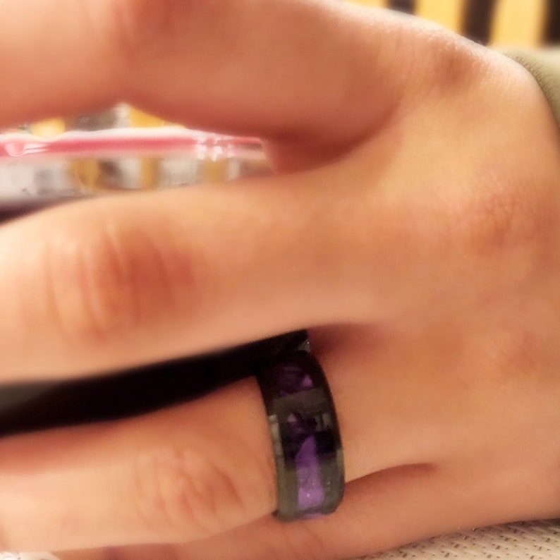Pode incluir: Um anel preto com um design de incrusta&ccedil;&atilde;o roxo. O anel &eacute; usado em um dedo.