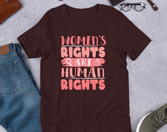 Könnte beinhalten: Ein maroonfarbenes T-Shirt mit dem Text "Women's Rights Are Human Rights" in rosa Schrift. Der Text ist mit einem rosa weiblichen Symbol und einem rosa Herzen mit einer Krone verziert.