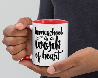 Puede incluir: Taza de cerámica blanca con borde rojo. La taza tiene texto negro que dice "homeschool is a work of heart" con un pequeño icono de sobre con un corazón dentro.