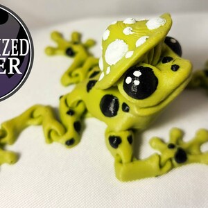 May include: A green frog figurine with black spots and a white mushroom hat. The frog has large black eyes and a wide smile.