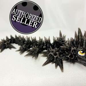 May include: A black, spiky, 3D printed dragon figurine with a yellow eye. The dragon is posed with its head turned to the right and its body curled slightly. The figurine is made of a textured material that gives it a rough, scaly appearance.