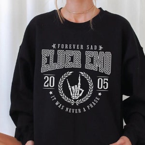 以下が含まれることがあります： 「Forever Sad Elder Emo 20 05 It Was Never A Phase」と白で書かれたグラフィックプリントが施された黒いスウェットシャツ。グラフィックには、ロックンロールサインをしているスケルトンの手も含まれています。