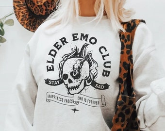 May include: A light gray sweatshirt with the text "ELDER EMO CLUB" and a skull graphic. The sweatshirt also features the phrases "STAY SAD" and "EMO IS FOREVER". A brown and black leopard print tote bag is visible.