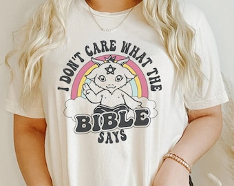 May include: A white t-shirt with a graphic of a cartoon goat wearing a crown and a star on its forehead. The goat is standing in front of a rainbow and the text "I don't care what the bible says" is printed on the shirt.