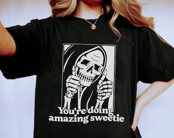 May include: Black t-shirt with a graphic of a skeleton wearing a hooded robe and giving a thumbs up. The text "You're doing amazing sweetie" is printed below the graphic.