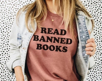 May include: A woman wearing a mauve t-shirt with the text "READ BANNED BOOKS" in black. She is wearing a denim jacket and black jeans.