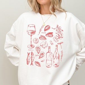 May include: White sweatshirt with a red illustration of wine and seafood. The design includes a wine glass, bottles, oysters, sardines, mussels, and a corkscrew. The words "sardines" and "oyster" are also included in the design.