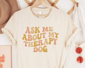 May include: A beige t-shirt with the text "Ask me about my therapy dog" in a retro font with pink, orange, and yellow colors.