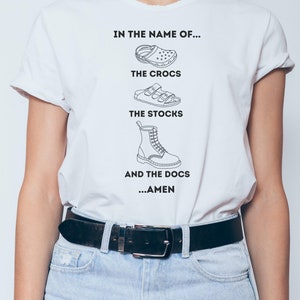 May include: White t-shirt with a graphic print of different types of shoes and the text "In the name of... The Crocs, The Stocks, And The Docs... Amen".