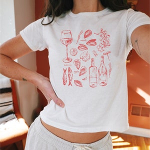 May include: White t-shirt featuring a red illustration of a glass of wine, two bottles of wine, a corkscrew, and various seafood including oysters, mussels, and sardines. The text "oysters", "mussels", and "sardines" is included in the illustration.