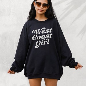 May include: Black sweatshirt with the text "West Coast Girl" in a white, retro font. The sweatshirt has long sleeves and a relaxed fit. The image shows a person wearing sunglasses.