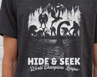 May include: A dark heather grey t-shirt with a white graphic print of various mythical creatures playing hide and seek in a forest. The text "Hide & Seek World Champions League" is printed below the graphic.