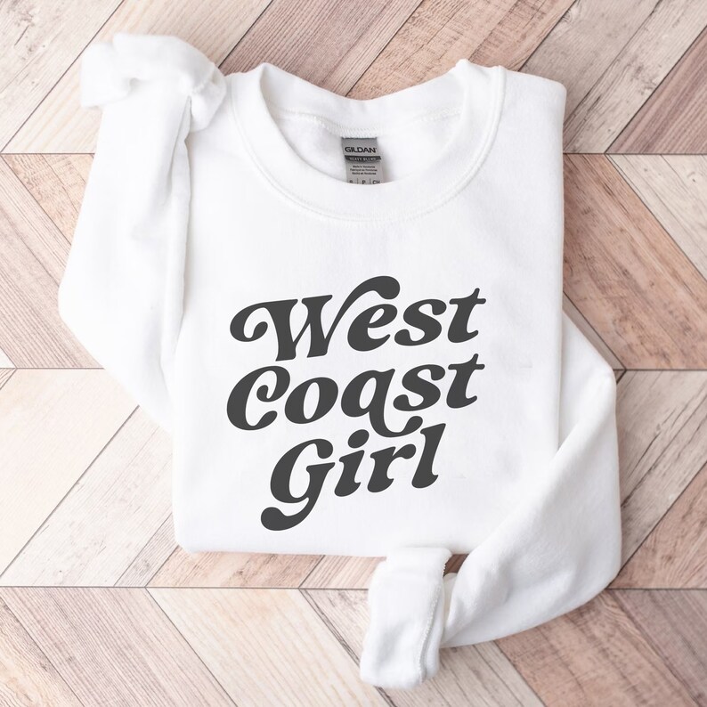May include: A white sweatshirt with the words "West Coast Girl" in a dark gray, retro font. The long-sleeved top is folded neatly on a light brown wooden surface. The Gildan brand label is visible at the back of the neck.