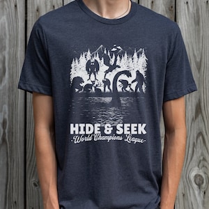 May include: A dark blue t-shirt with a white graphic of various mythical creatures hiding in a forest. The text "HIDE & SEEK. World Champions League." is printed below the graphic.