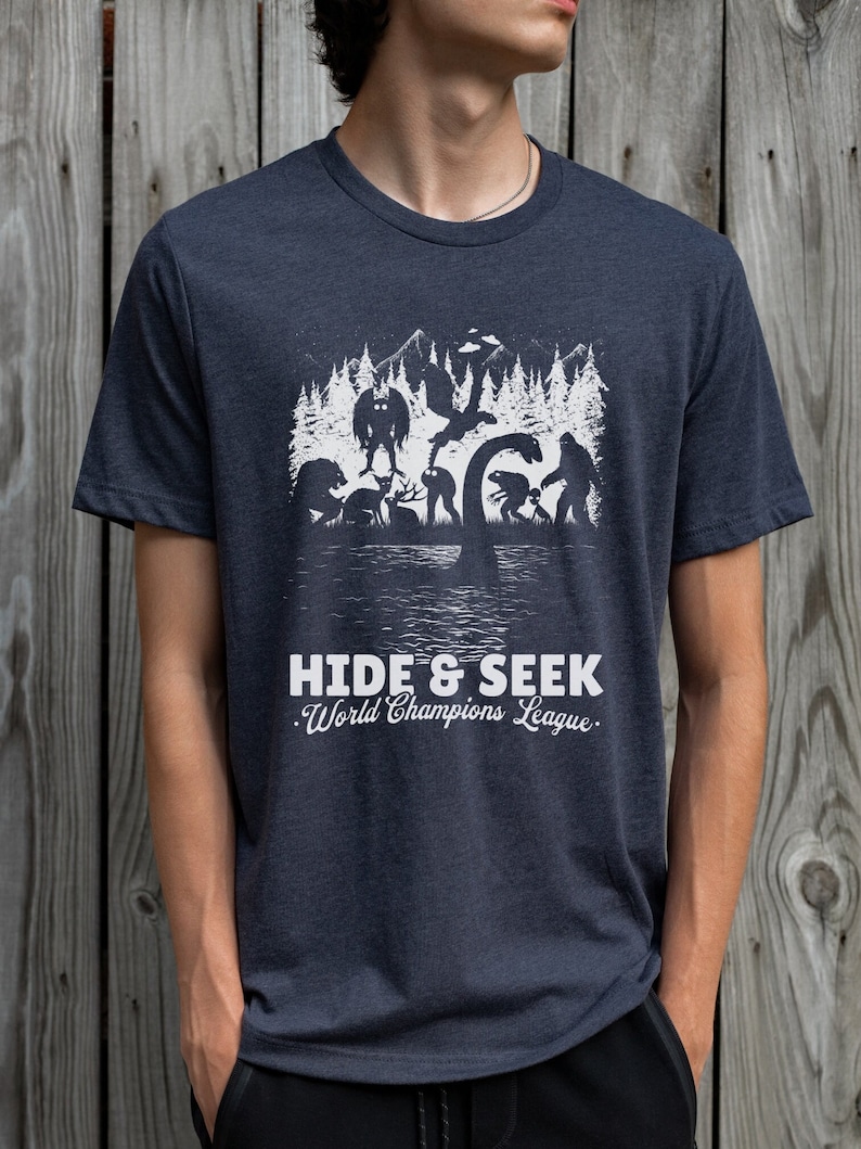 May include: A dark blue t-shirt with a white graphic of various mythical creatures hiding in a forest. The text "HIDE & SEEK. World Champions League." is printed below the graphic.