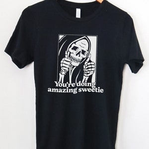 May include: Black t-shirt featuring a white graphic of a skeleton in a hooded robe giving a thumbs up. The text "You're doing amazing sweetie" is printed below the graphic.