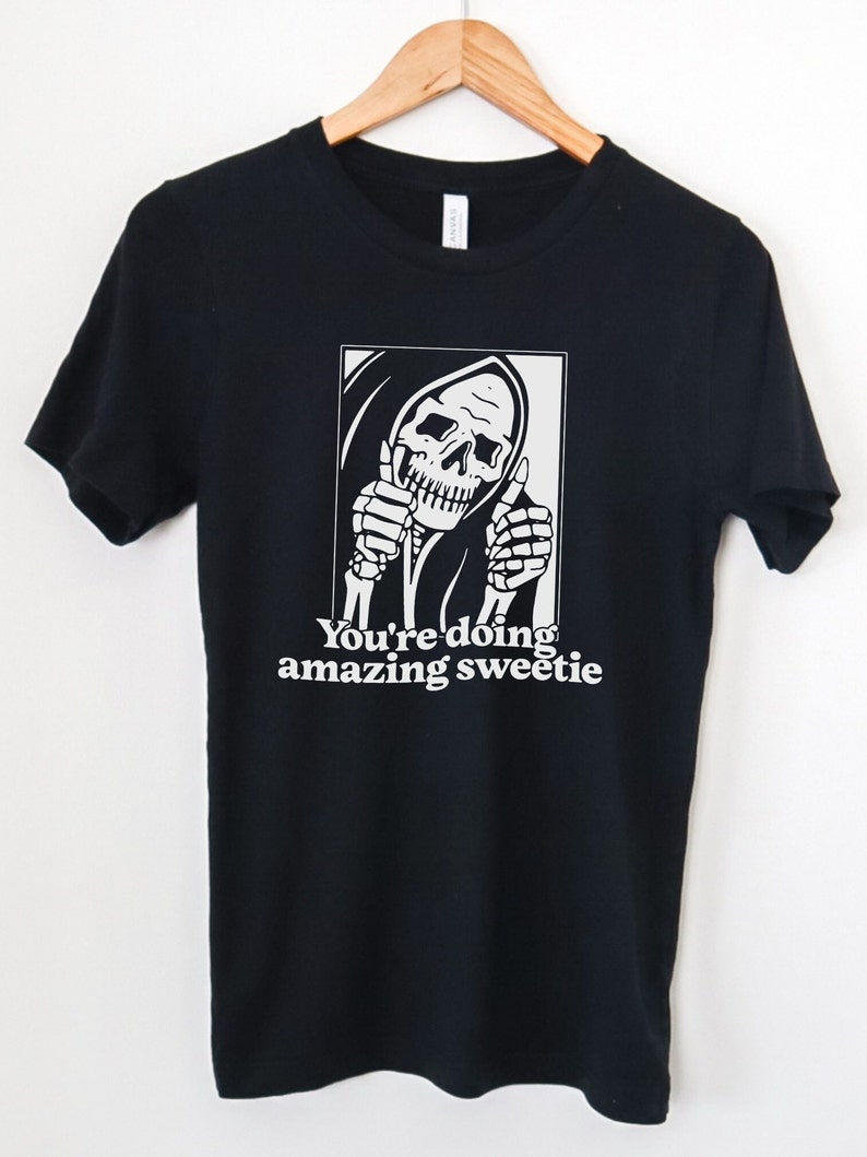 May include: Black t-shirt featuring a white graphic of a skeleton in a hooded robe giving a thumbs up. The text "You're doing amazing sweetie" is printed below the graphic.