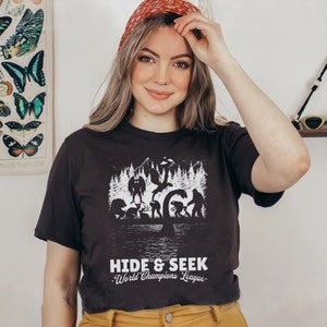 May include: A black t-shirt with a white graphic of various mythical creatures in a forest setting. The text "HIDE & SEEK World Champions League." is printed below the graphic.