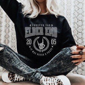 以下が含まれることがあります： 「Forever Sad Elder Emo 20 05 It Was Never A Phase」と白文字で書かれた黒いスウェットシャツ。テキストは市松模様で、花輪で囲まれています。花輪の中央には、ロックンロールのハンドジェスチャーをしたスケルトンの手があります。