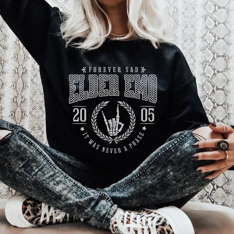 以下が含まれることがあります： 「Forever Sad Elder Emo 20 05 It Was Never A Phase」と白文字で書かれた黒いスウェットシャツ。テキストは市松模様で、花輪で囲まれています。花輪の中央には、ロックンロールのハンドジェスチャーをしたスケルトンの手があります。