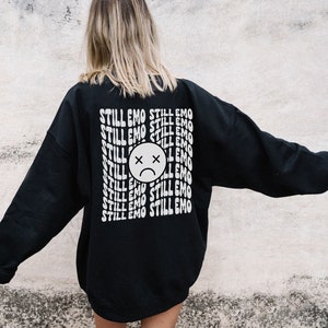 May include: Black sweatshirt with a white graphic print of a sad face and the text "STILL EMO" repeated multiple times.
