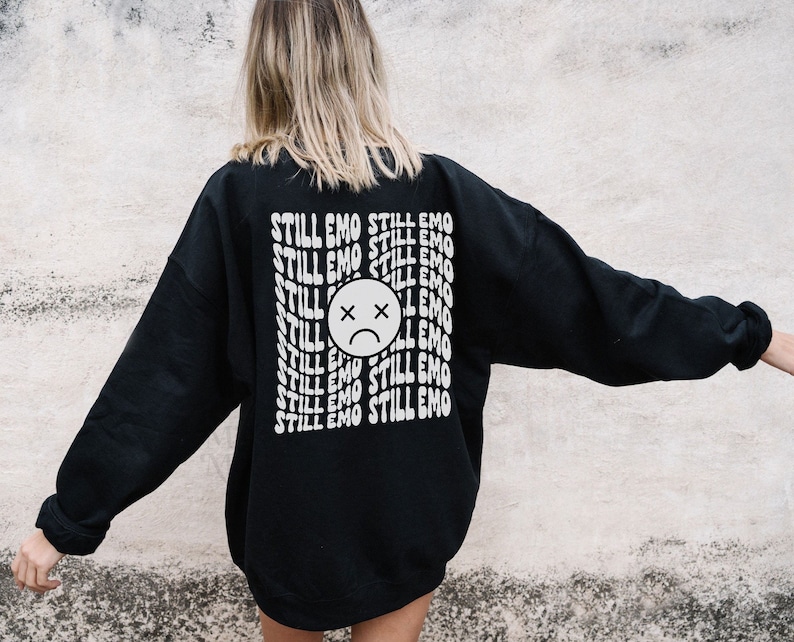 May include: Black sweatshirt with a white graphic print of a sad face and the text "STILL EMO" repeated multiple times.