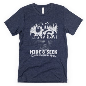 May include: A navy blue t-shirt with a white graphic of a group of mythical creatures playing hide and seek in a forest. The text "Hide & Seek World Champions League" is printed below the graphic.