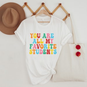 May include: White t-shirt with the text "YOU ARE ALL MY FAVORITE STUDENTS" in rainbow colours.