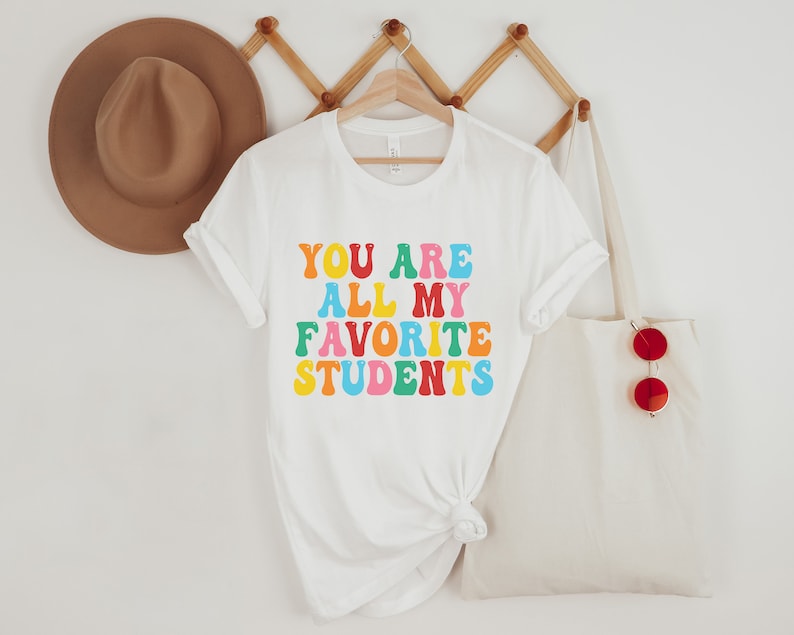May include: White t-shirt with the text "YOU ARE ALL MY FAVORITE STUDENTS" in rainbow colours.